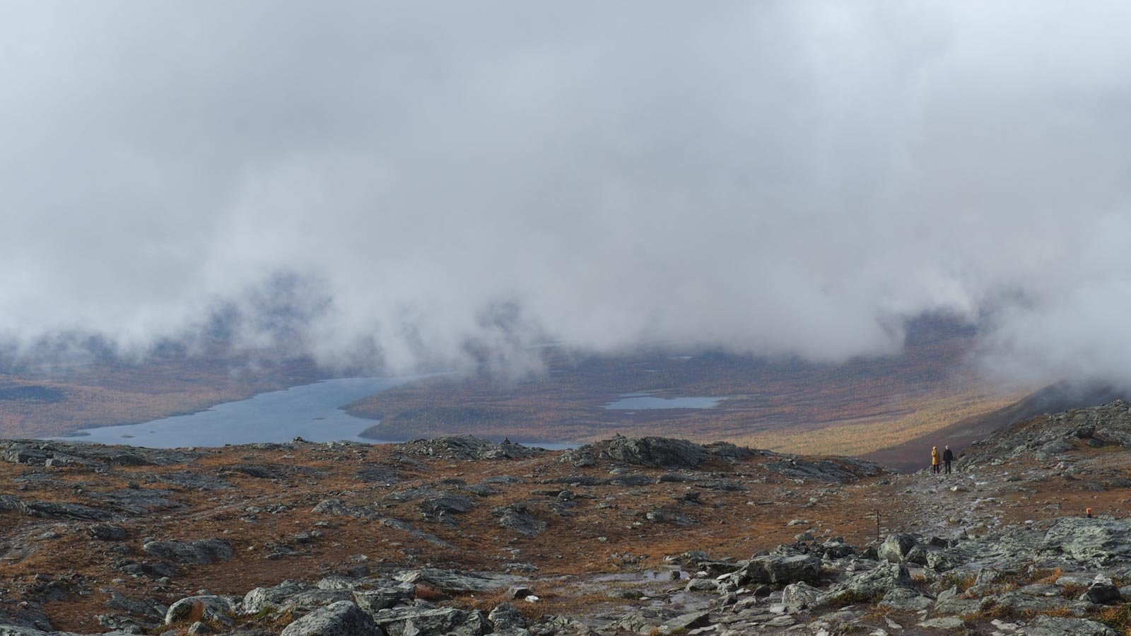 Näkymä Saanatunturin laelta: etualalla näkyy kivistä tunturin lakea, mutta matalalla roikkuvat pilvet peittävät näkymän laaksoon. 