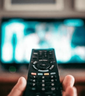Close-up of remote control pointed at blurred TV screen in background